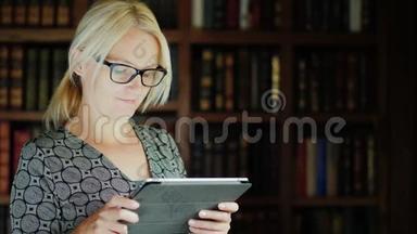 一个戴眼镜的漂亮女人在图书馆里用平板电脑。 站在书架和书的背景下